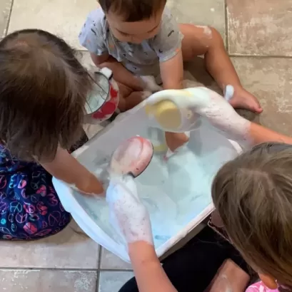 two toddlers and an older child sitting on the floor around a white wash bin filled with bubble foan. They are playing with the foam and using bowls and cups mixing and pouring the foam inside the container.