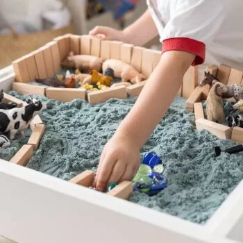 child plaing with gray sand in sensory bin with blocks and farm animals